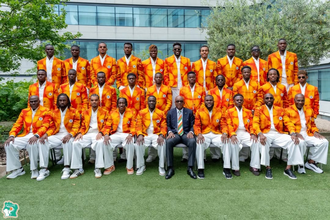 The complete Côte d'Ivoire national football squad posing outdoors on grass for an official World Cup team photograph wearing matching orange Ibrahim Fernandez tie-dye blazers and white trousers.