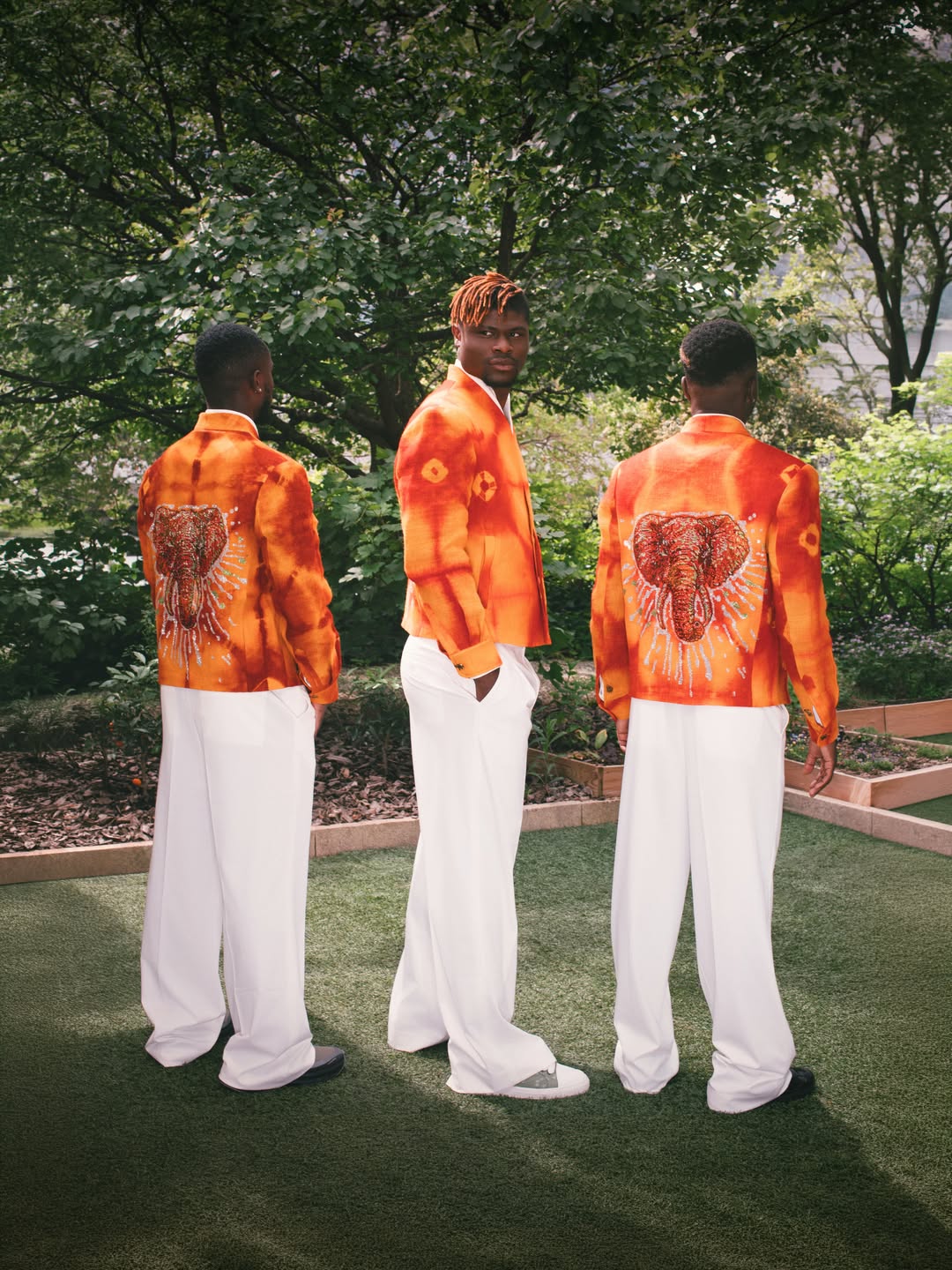 Three Côte d'Ivoire football players in an outdoor garden showcasing the backs of their orange tie-dye blazers designed by Ibrahim Fernandez, featuring a large embroidered elephant emblem.