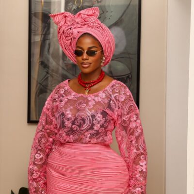 Portrait of Hilda Baci highlighting her sheer pink floral lace buba bodice, chunky red coral bead necklace, and retro sunglasses, styled by Emmanuel Goodnews.