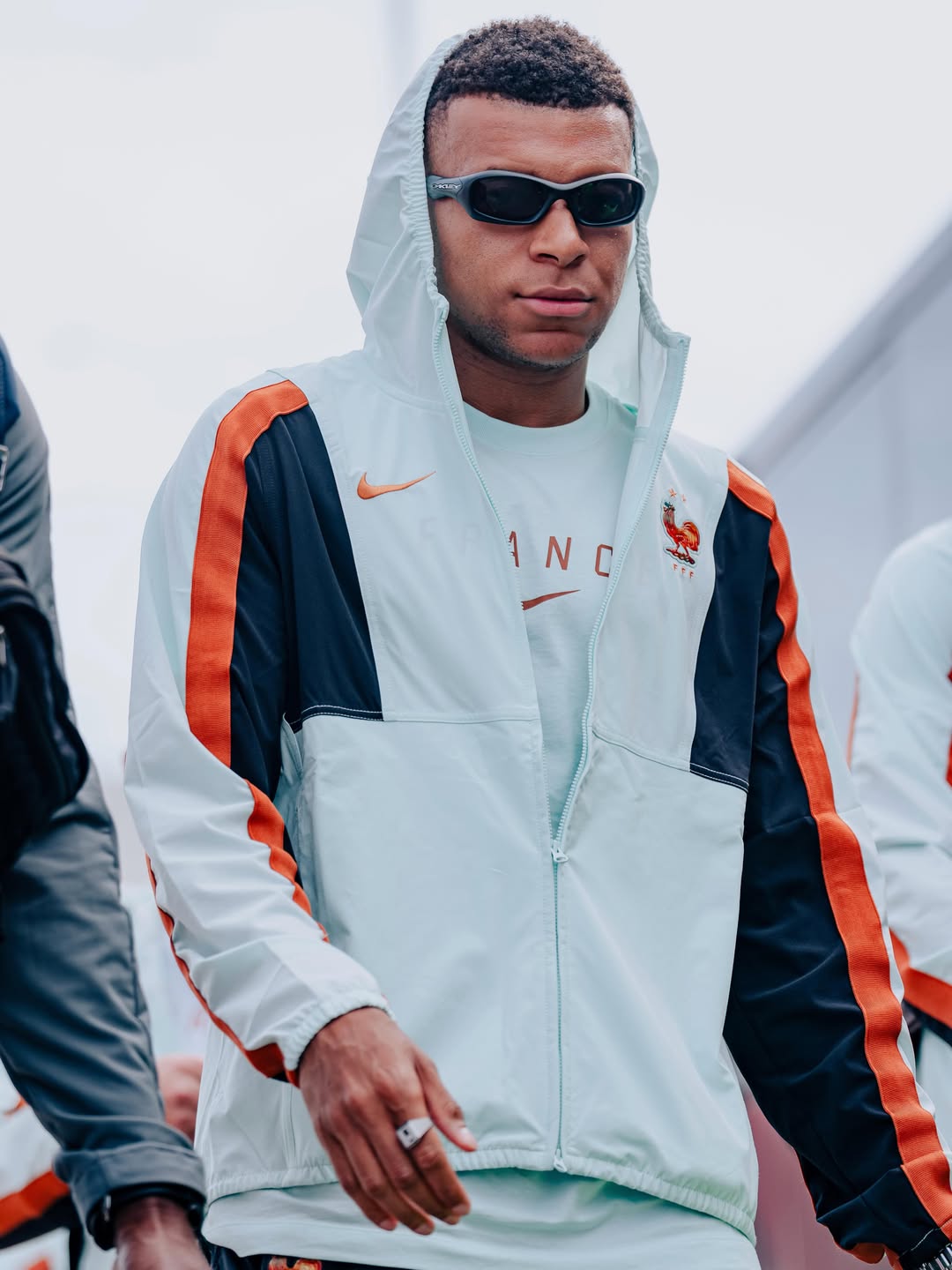 Kylian Mbappe wearing sunglasses and a hooded white and blue France national team tracksuit while walking outdoors upon arriving in the US.