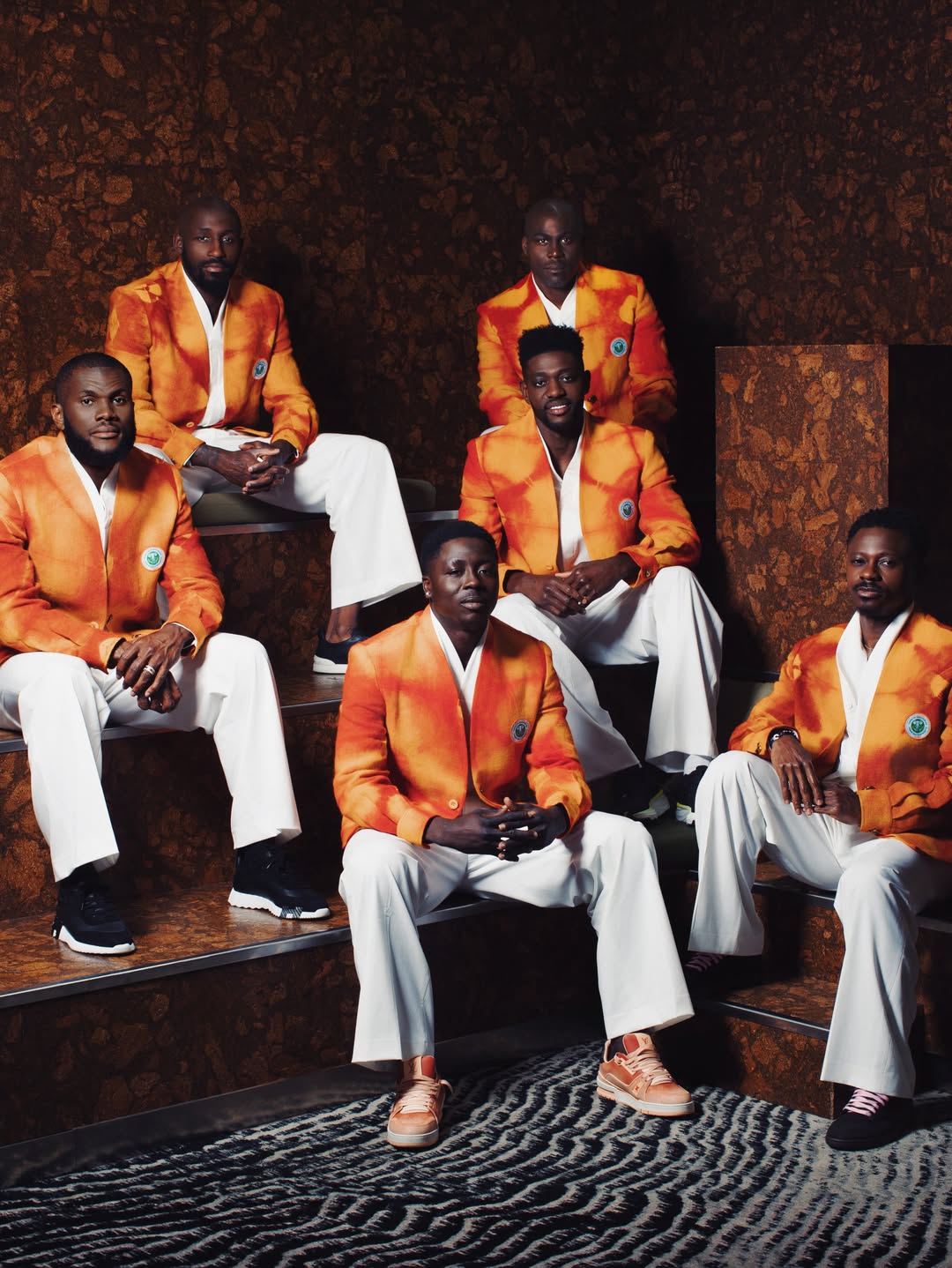 Six members of the Côte d’Ivoire football squad posing together on carpeted steps in matching orange tie-dye luxury blazers by Ibrahim Fernandez and white trousers.