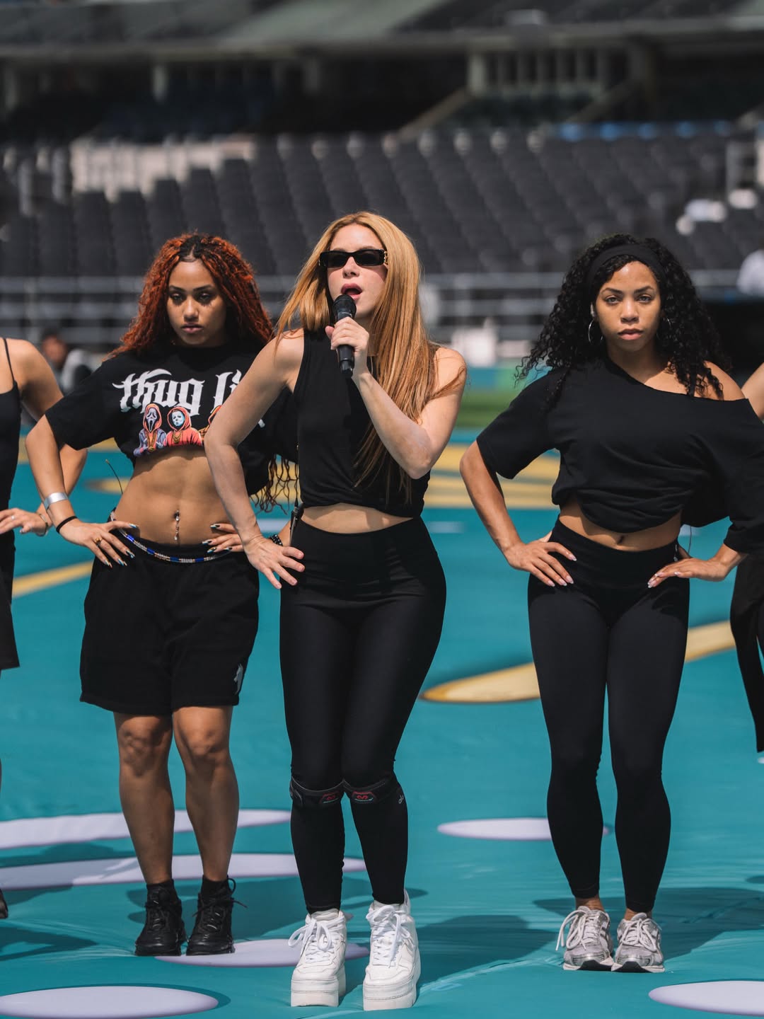 Shakira in a black top and black leggings holding a microphone during an outdoor stadium choreography rehearsal with two dancers.