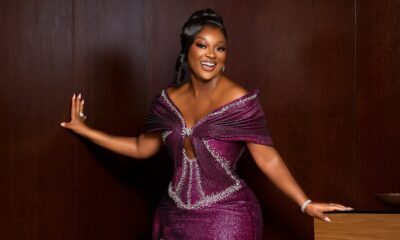 Full-length portrait of Ghanaian actress Jackie Appiah posing against a dark wood wall in a custom metallic magenta column gown with structured fan pleating.