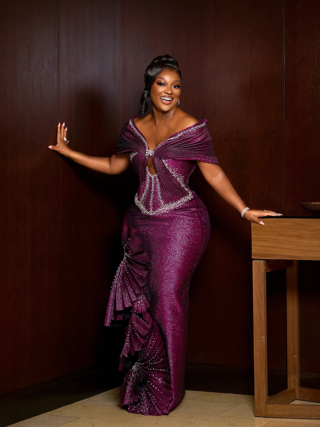 Full-length portrait of Ghanaian actress Jackie Appiah posing against a dark wood wall in a custom metallic magenta column gown with structured fan pleating.