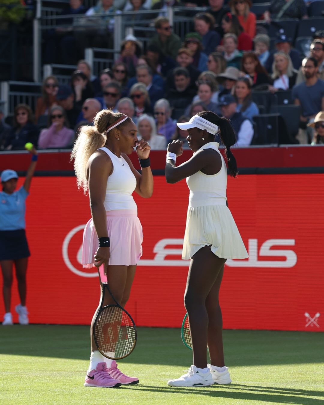 Serena Williams and Victoria Mboko standing close together on court holding their rackets and discussing tactics during their doubles debut.