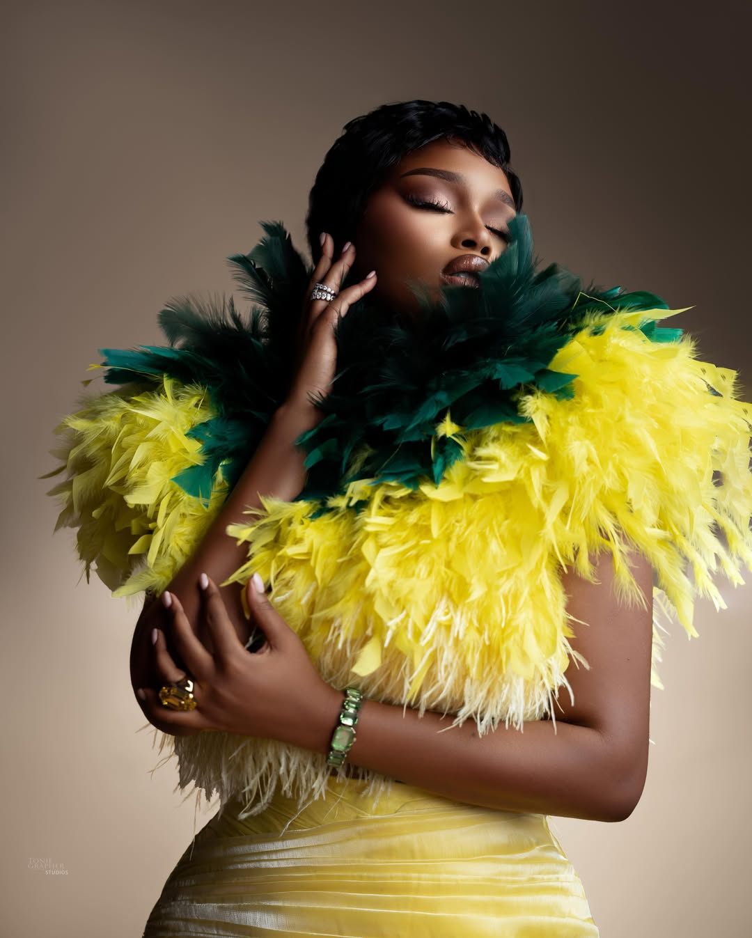 Sabrina Idukpaye framed by layers of yellow and forest green feathers with a soft smoky eye makeup look.