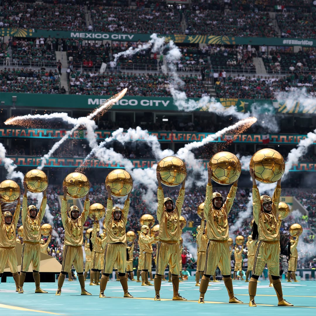 Performers dressed in metallic gold suits lifting large golden footballs under stadium smoke trails.