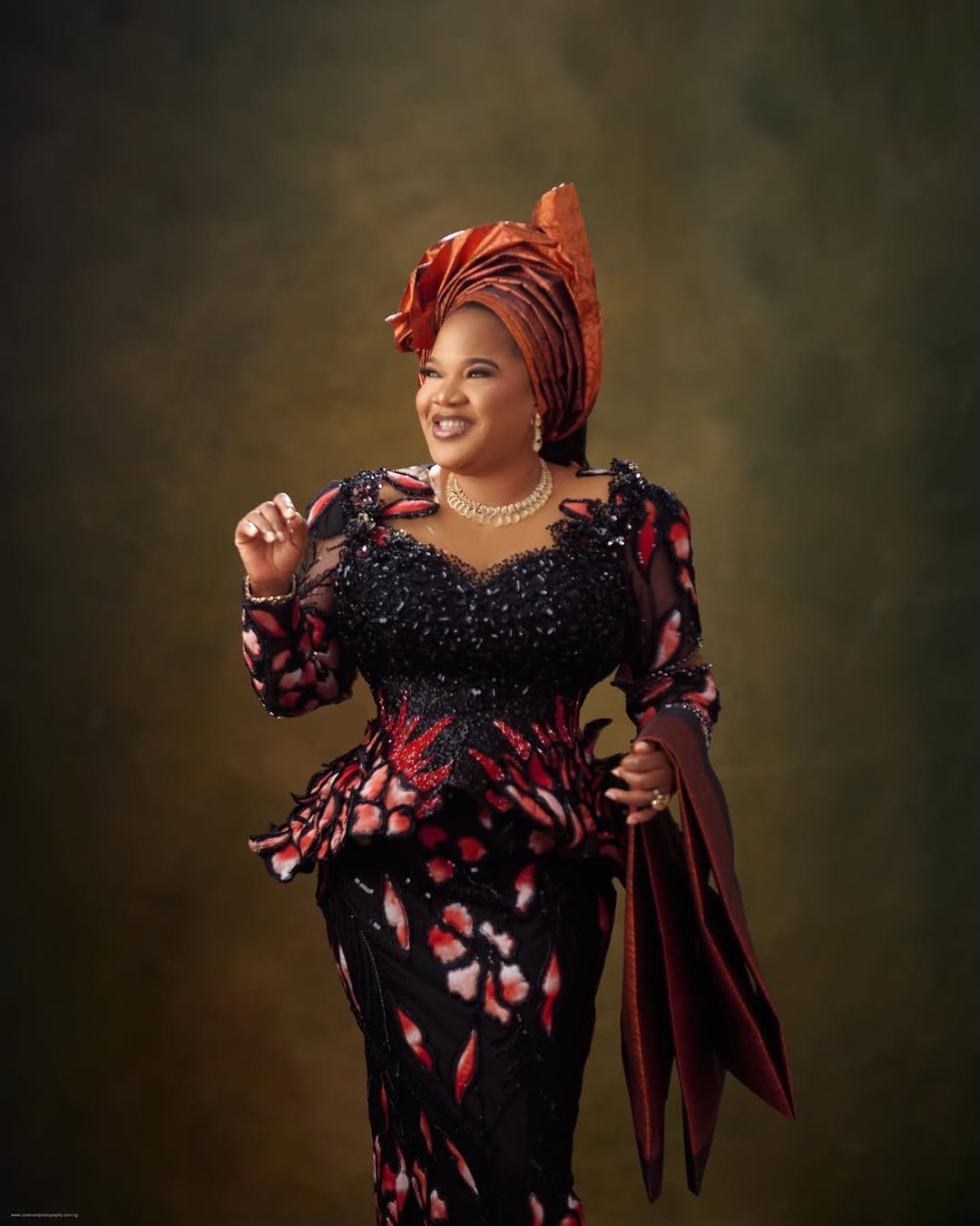 Toyin Abraham Ajeyemi smiling in a black and terracotta beaded lace peplum blouse holding a matching shoulder cloth.