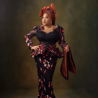 Toyin Abraham Ajeyemi posing with one hand on her hip, wearing a pleated copper architectural gele, a terracotta beaded lace outfit, and holding a matching ipele over her arm.