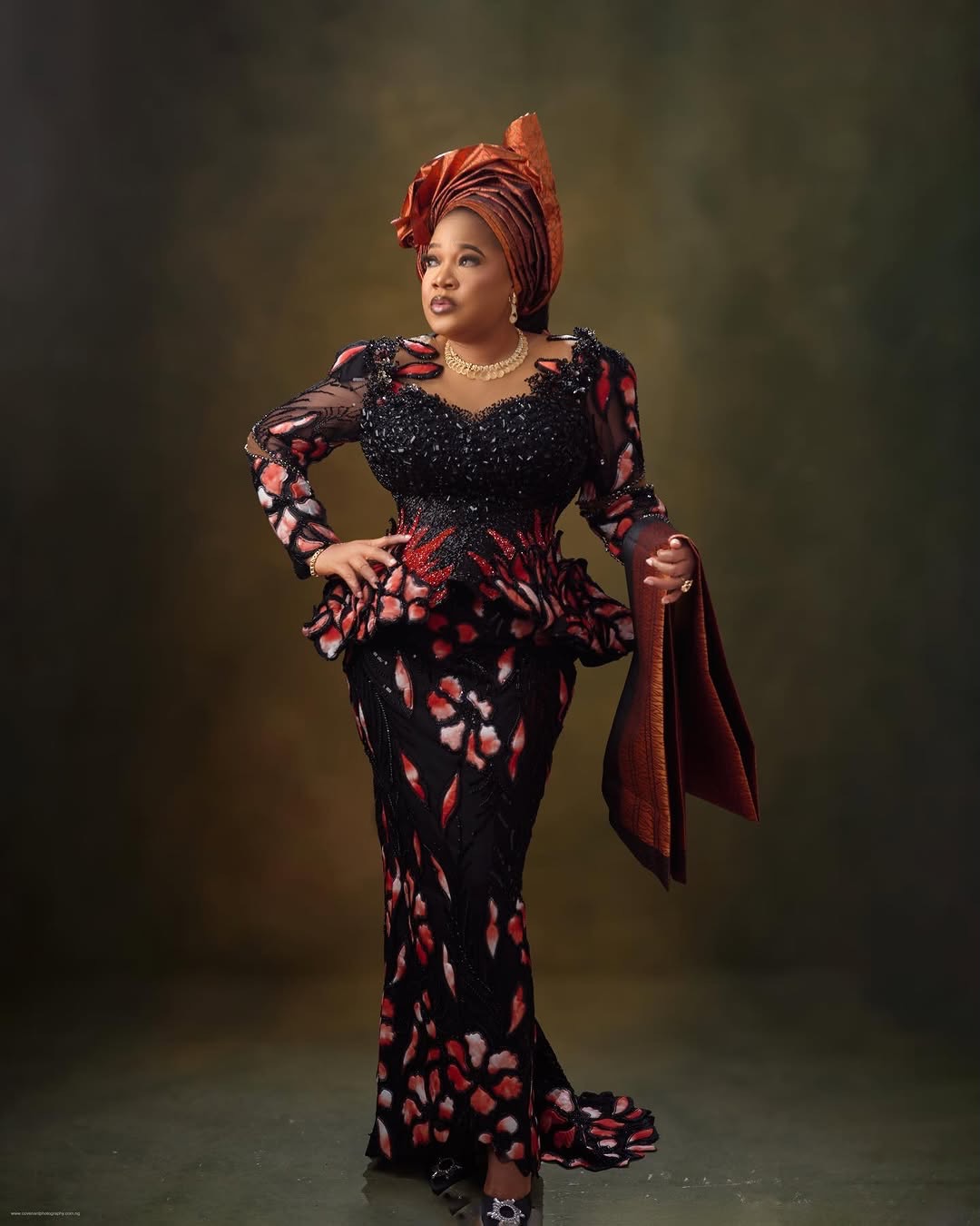 Toyin Abraham Ajeyemi posing with one hand on her hip, wearing a pleated copper architectural gele, a terracotta beaded lace outfit, and holding a matching ipele over her arm.