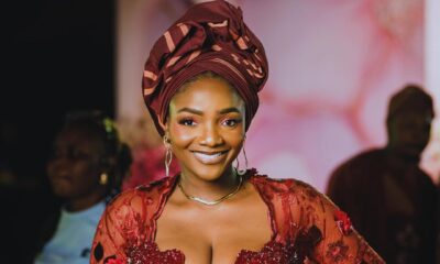 Simi smiling broadly in a heavily embellished wine-coloured lace outfit with gold hoop earrings and matching red nails.
