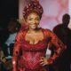 Simi smiling broadly in a heavily embellished wine-coloured lace outfit with gold hoop earrings and matching red nails.
