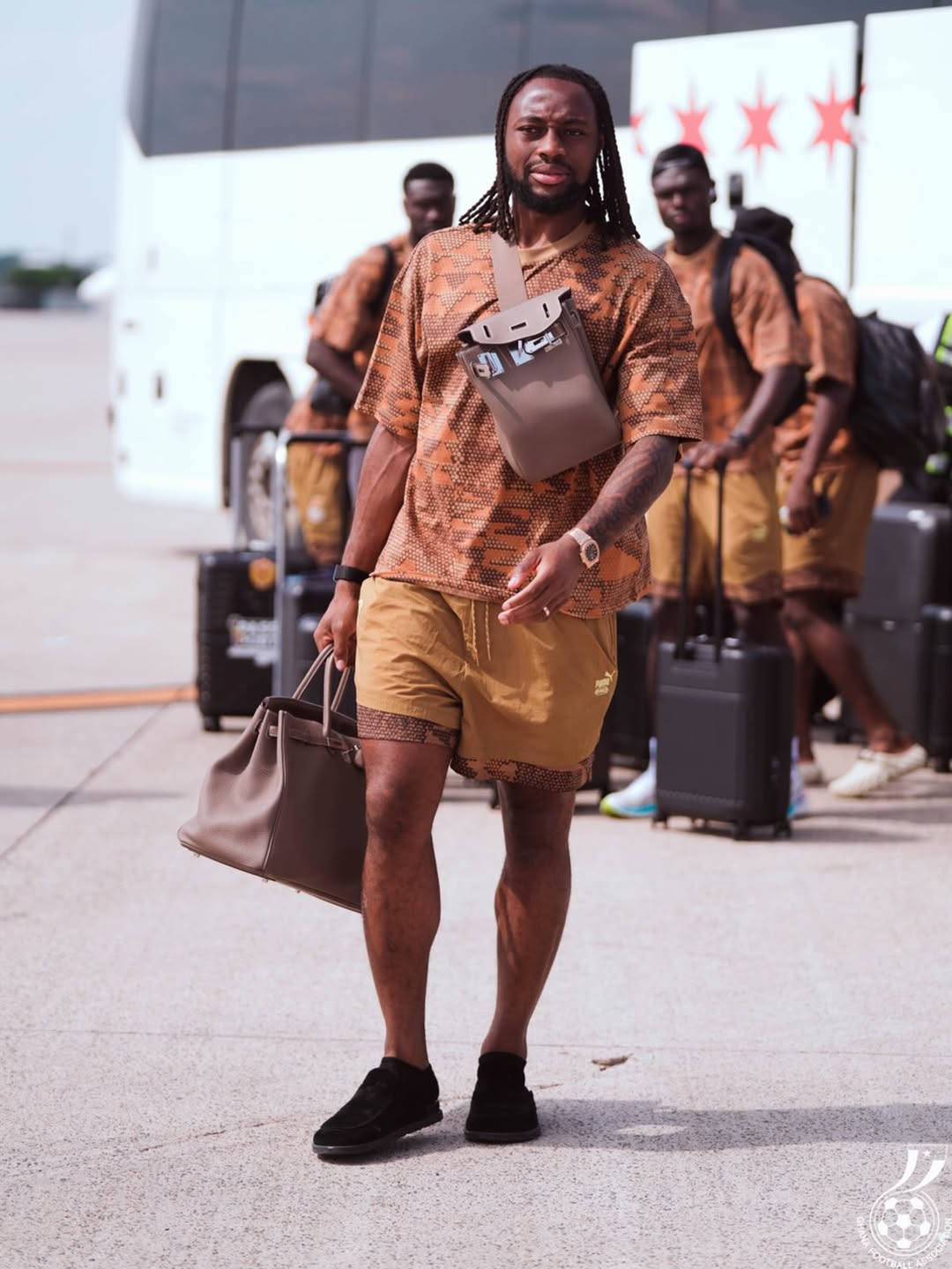 Ghana forward Antoine Semenyo walking on the airport tarmac in Canada carrying a brown leather travel bag and wearing a patterned brown short-sleeve shirt and shorts set.