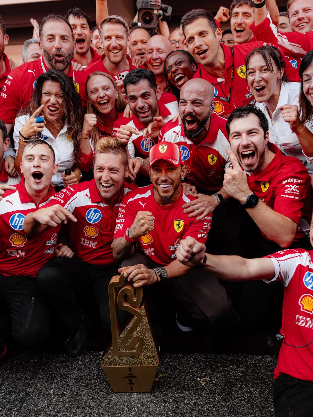 Lewis Hamilton smiles and holds his golden race trophy while posing for a group picture with the Scuderia Ferrari crew.