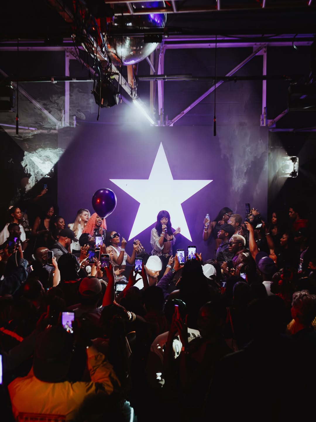 A wide shot of Ayra Starr performing on a dimly lit venue stage in front of a massive illuminated white star backdrop while fans hold up phones.