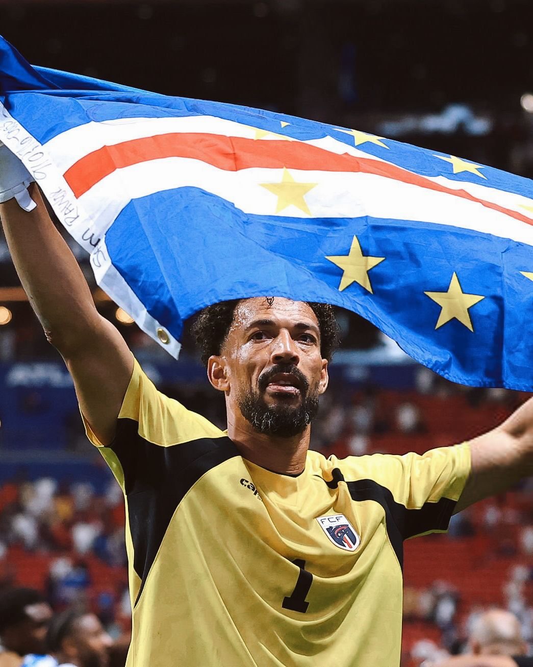 Goalkeeper Vozinha smiling and holding the blue, white, and red national flag of Cabo Verde above his head in the stadium.