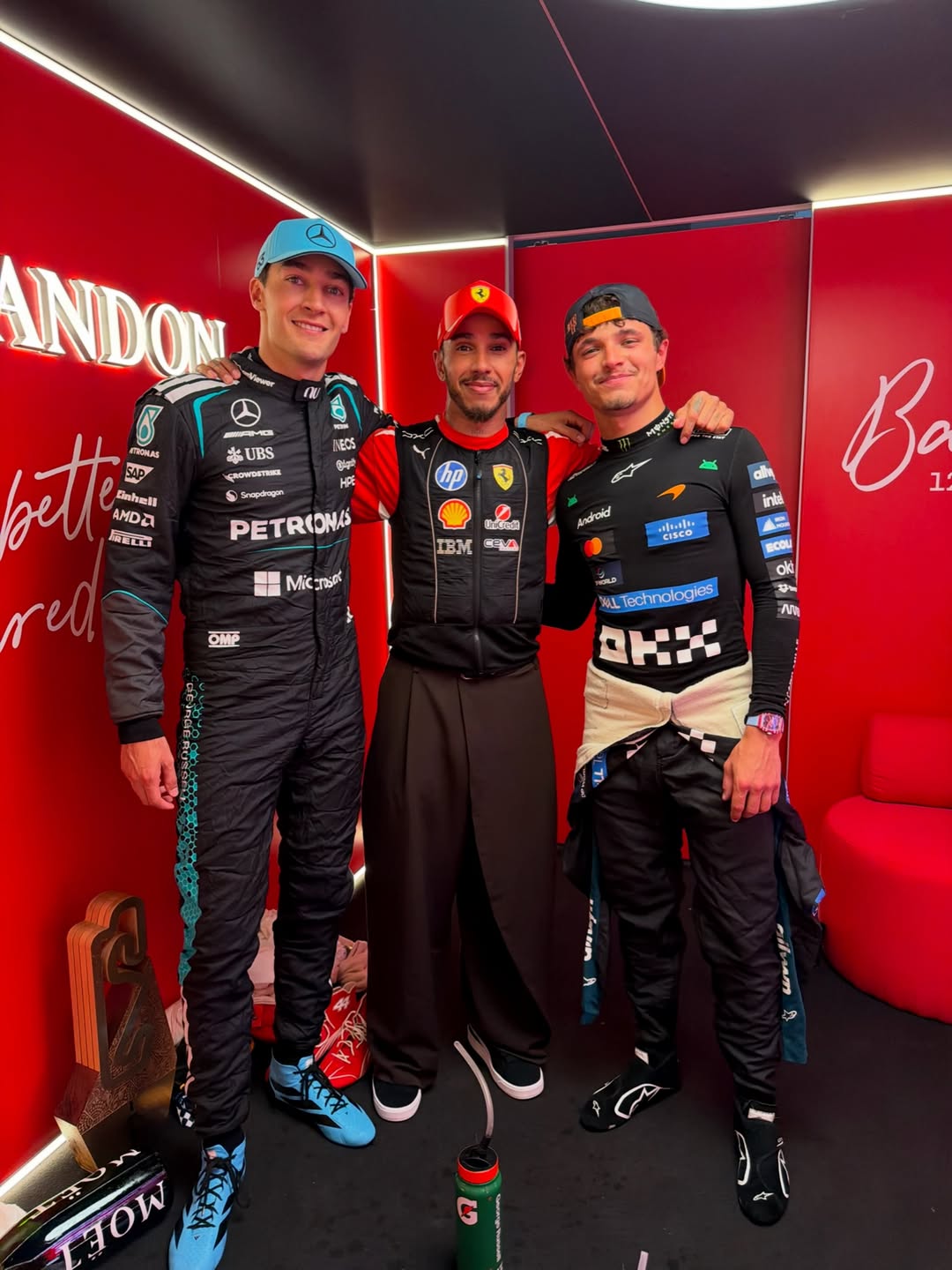 Formula 1 drivers George Russell, Lewis Hamilton, and Lando Norris stand together in racing suits inside a red room.
