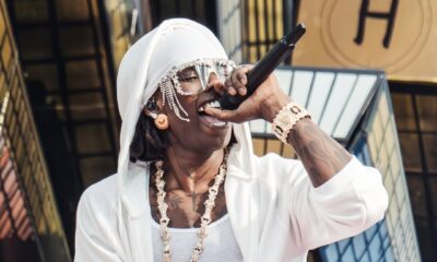 Rema holds a microphone to his mouth while wearing a white hooded jacket, a matched white wrap, and his angel chain pendant.
