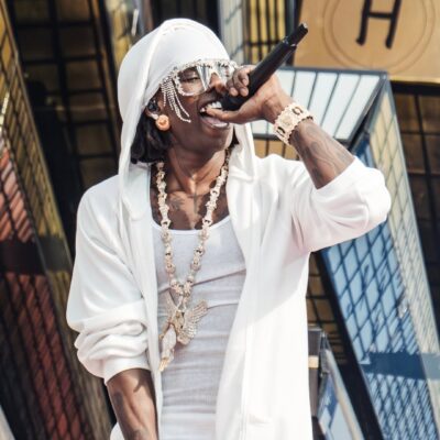 Rema holds a microphone to his mouth while wearing a white hooded jacket, a matched white wrap, and his angel chain pendant.