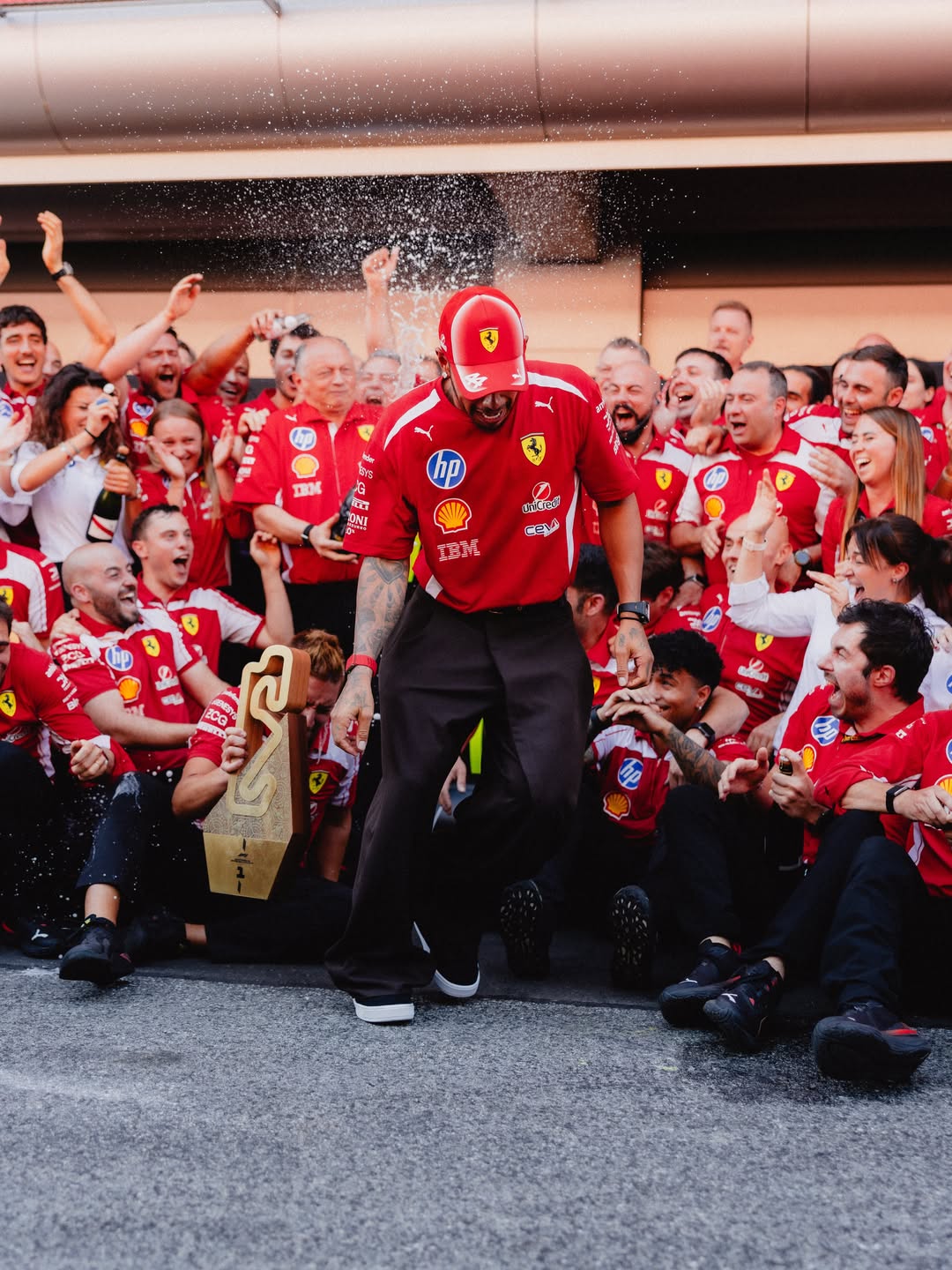 Formula 1 driver Lewis Hamilton in a red Ferrari kit celebrates on the ground surrounded by cheering Scuderia Ferrari team members.