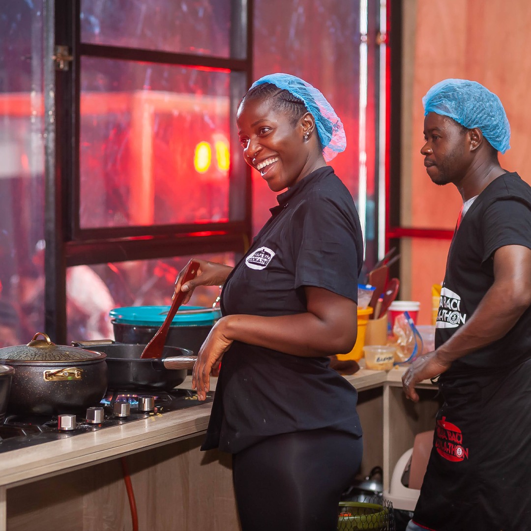 Hilda Baci wearing a black cook-a-thon shirt and a blue hairnet smiling broadly while using a wooden spoon to stir a pan over a lit gas stove burner during her first record attempt in Lagos.