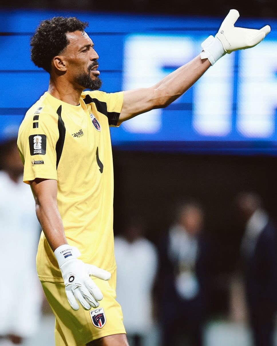 Cabo Verde goalkeeper Vozinha wearing white gloves and a yellow jersey while raising his arm to direct his team on the pitch.