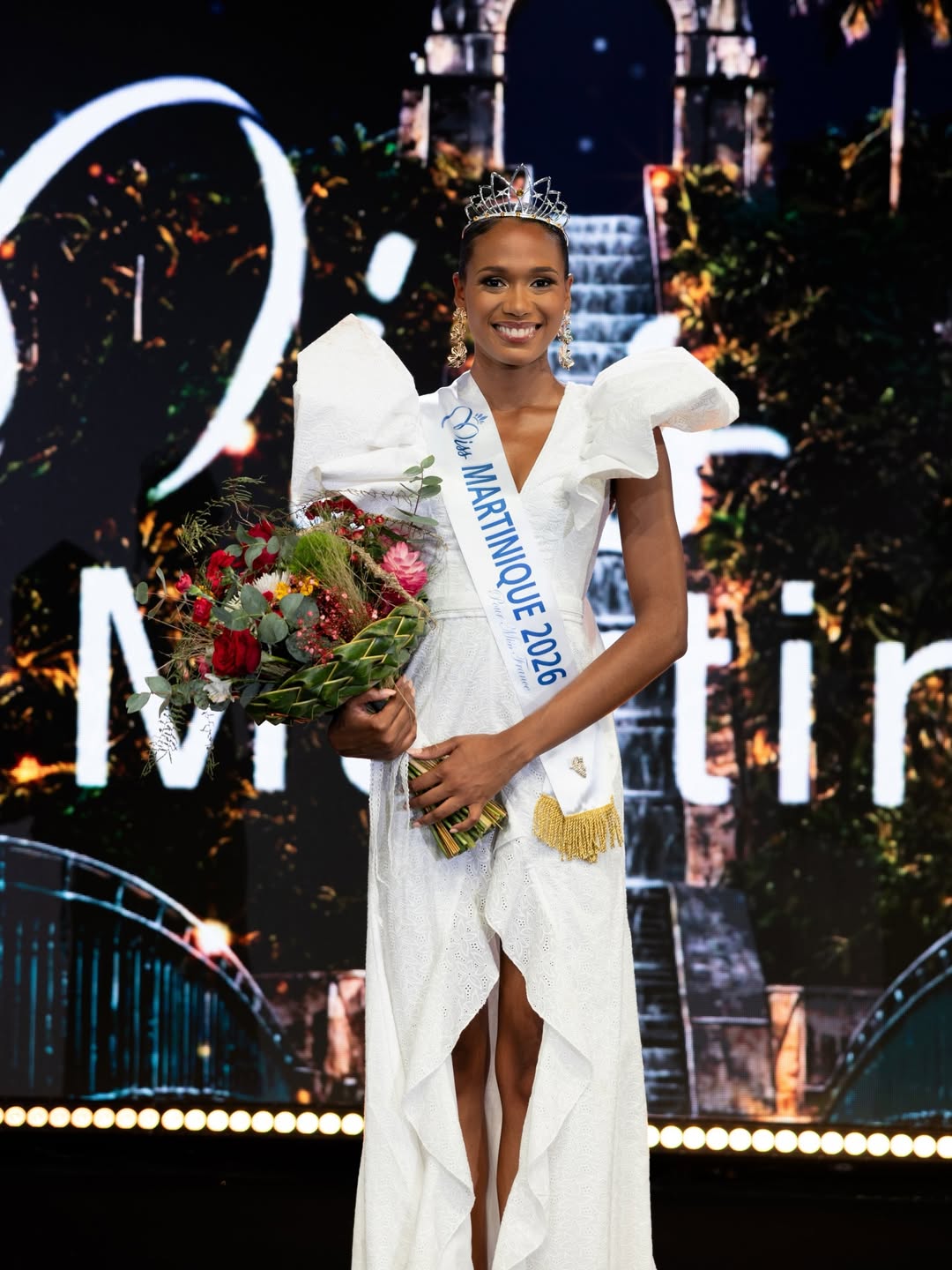 Miss Martinique 2026, Maureen-Alycia Lucéa-Merlin, smiling on stage in a white dress, tiara, and official sash while holding a colourful flower bouquet.