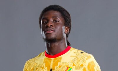 Studio portrait of Ghana national team football player Caleb Marfo Yirenkyi holding a FIFA match ball while wearing the official yellow and green patterned kit.