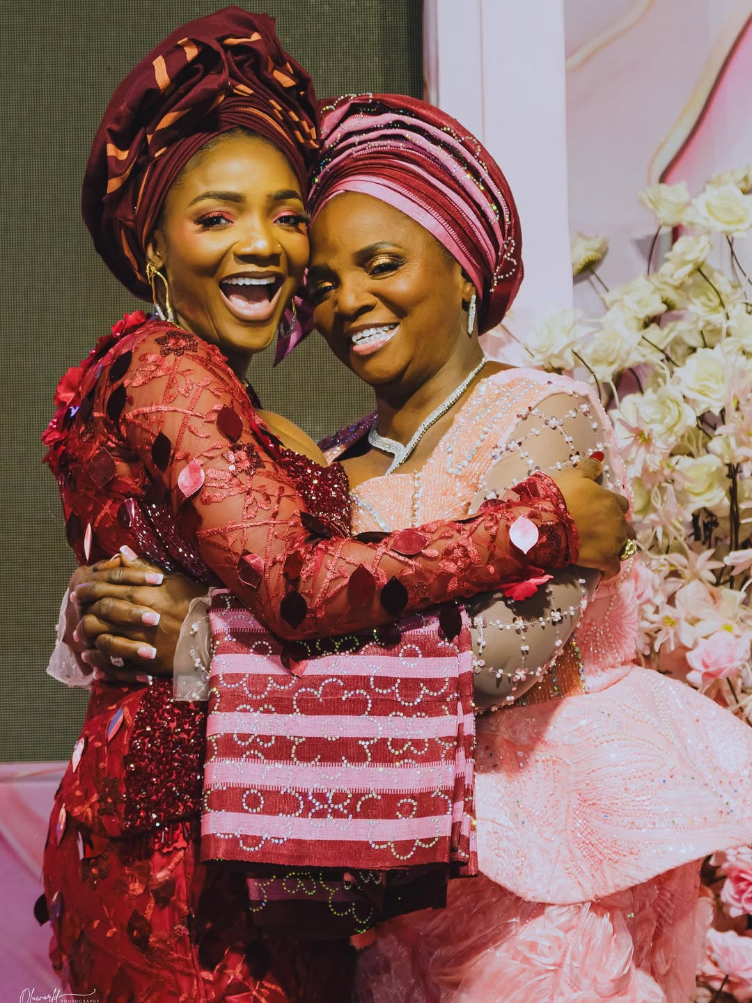 Simi in a burgundy traditional lace outfit hugging her mother Jedidiah Ogunleye who is wearing a light pink embellished gown at her 70th birthday party.