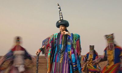 Denrele Edun standing in a desert landscape wearing a voluminous robe made of multi-coloured, vertically striped woven Aso-Oke with raw fringes, a towering black cultural headpiece, and heavy beaded necklaces while leaning on a wooden staff.