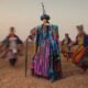 Denrele Edun standing in a desert landscape wearing a voluminous robe made of multi-coloured, vertically striped woven Aso-Oke with raw fringes, a towering black cultural headpiece, and heavy beaded necklaces while leaning on a wooden staff.