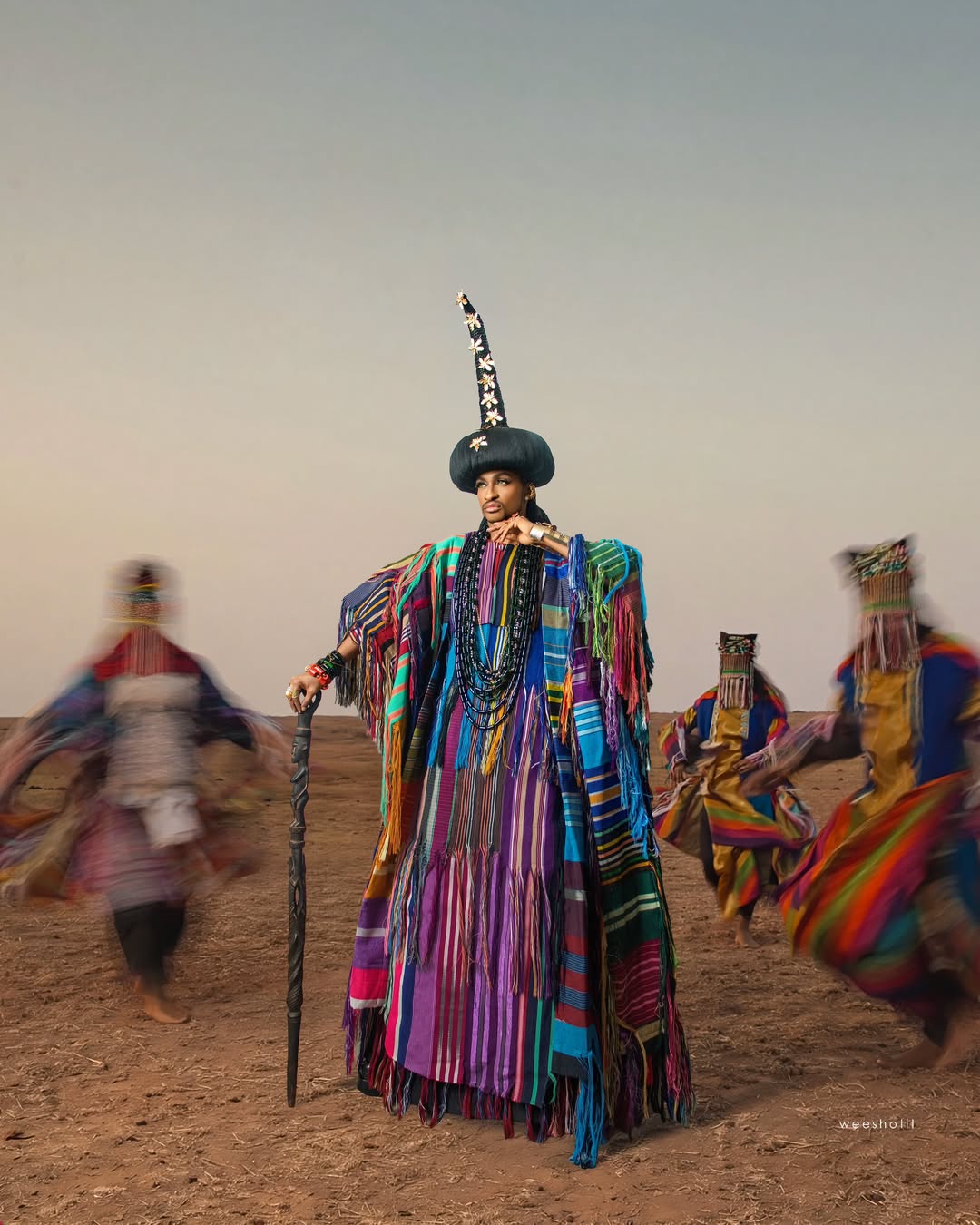 Denrele Edun standing in a desert landscape wearing a voluminous robe made of multi-coloured, vertically striped woven Aso-Oke with raw fringes, a towering black cultural headpiece, and heavy beaded necklaces while leaning on a wooden staff.