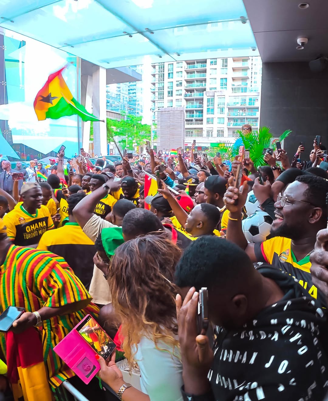 A large crowd of passionate Ghanaian football fans gathered in Toronto wearing yellow, red, and green team jerseys, waving the national flag of Ghana, and holding up miniature footballs to welcome the Black Stars