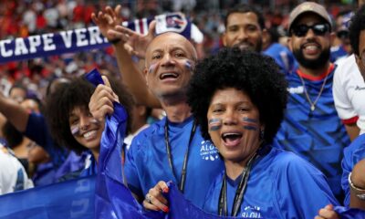 Happy Cabo Verde supporters with face paint wearing blue shirts and waving flags in the stadium stands.