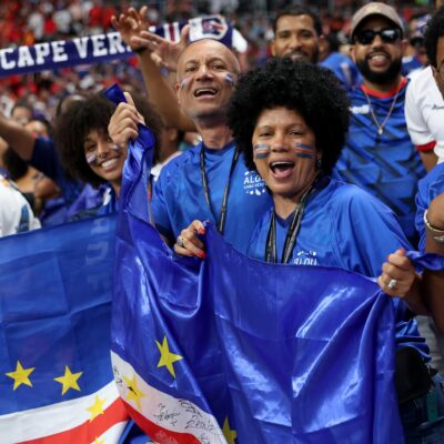 Happy Cabo Verde supporters with face paint wearing blue shirts and waving flags in the stadium stands.