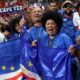 Happy Cabo Verde supporters with face paint wearing blue shirts and waving flags in the stadium stands.