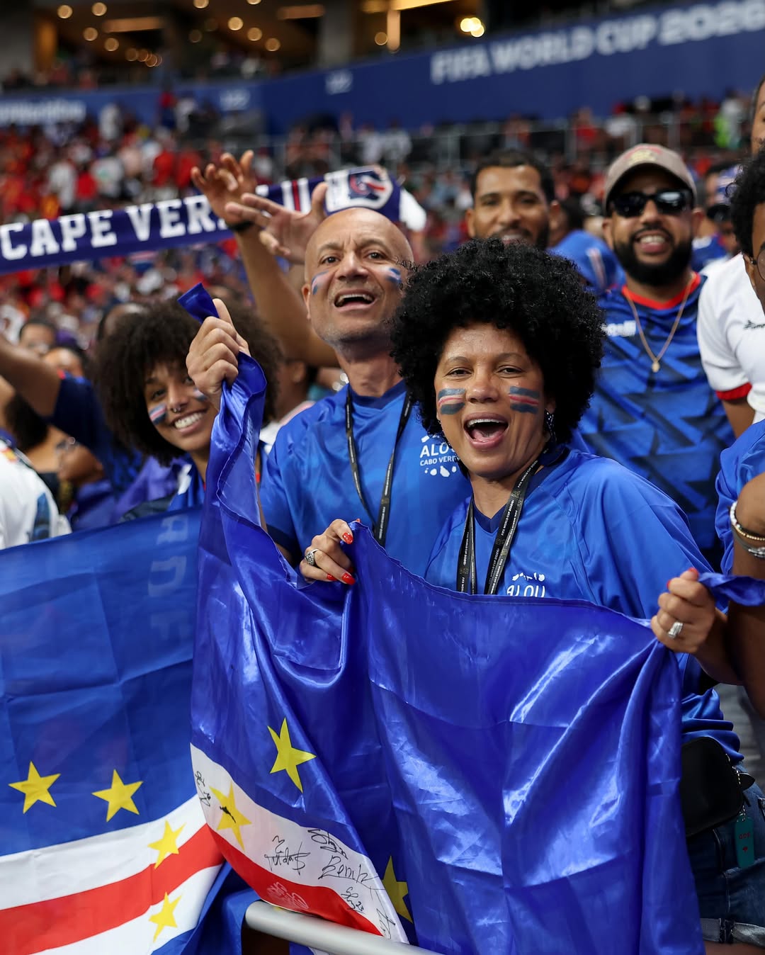 Happy Cabo Verde supporters with face paint wearing blue shirts and waving flags in the stadium stands.