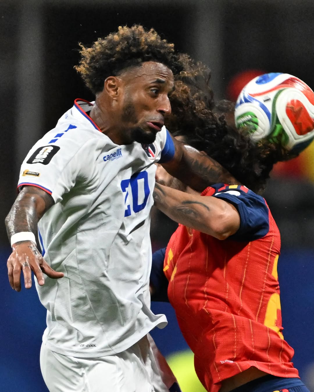 A Cabo Verde player in a white jersey competing in a tight aerial header duel against a Spanish player in a red jersey.