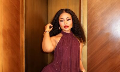Nigerian actress Teniola Aladese posing in an elevator alcove, wearing a shimmering burgundy halter mini dress and holding a matching leather mini cylinder bag.