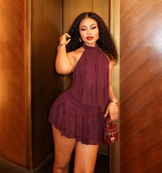 Nigerian actress Teniola Aladese posing in an elevator alcove, wearing a shimmering burgundy halter mini dress and holding a matching leather mini cylinder bag.