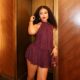 Nigerian actress Teniola Aladese posing in an elevator alcove, wearing a shimmering burgundy halter mini dress and holding a matching leather mini cylinder bag.