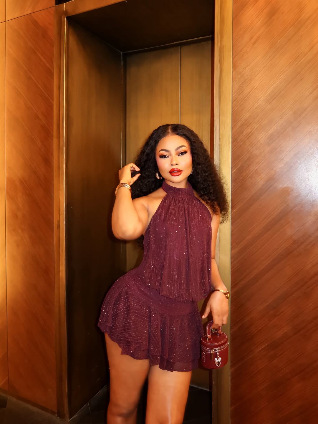 Nigerian actress Teniola Aladese posing in an elevator alcove, wearing a shimmering burgundy halter mini dress and holding a matching leather mini cylinder bag.