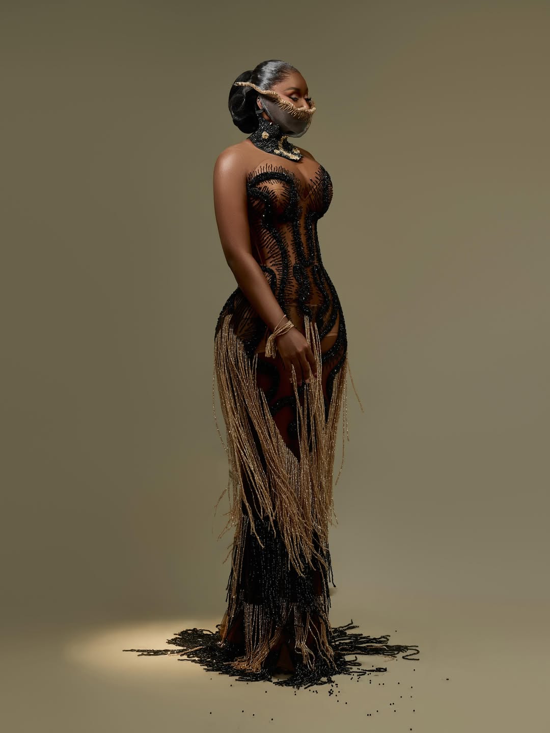 Nelly Mbonu wearing a sheer illusion birthday dress by Mub Rik embellished with vertical black beadwork, floor-length gold micro-bead fringe, and a custom metallic jawpiece.