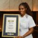 A portrait of Nigerian culinary star Hilda Baci in a white chef top presenting her official framed Guinness World Records Icon certificate.