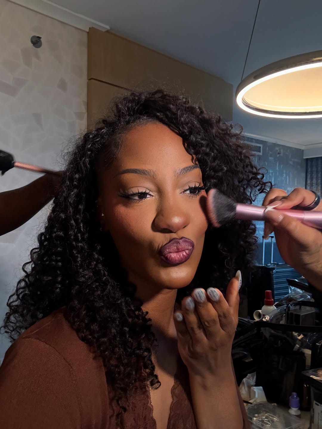 Tems getting her makeup done behind the scenes, featuring deep purple ombre lipstick, defined eyeliner, and voluminous dark curls.
