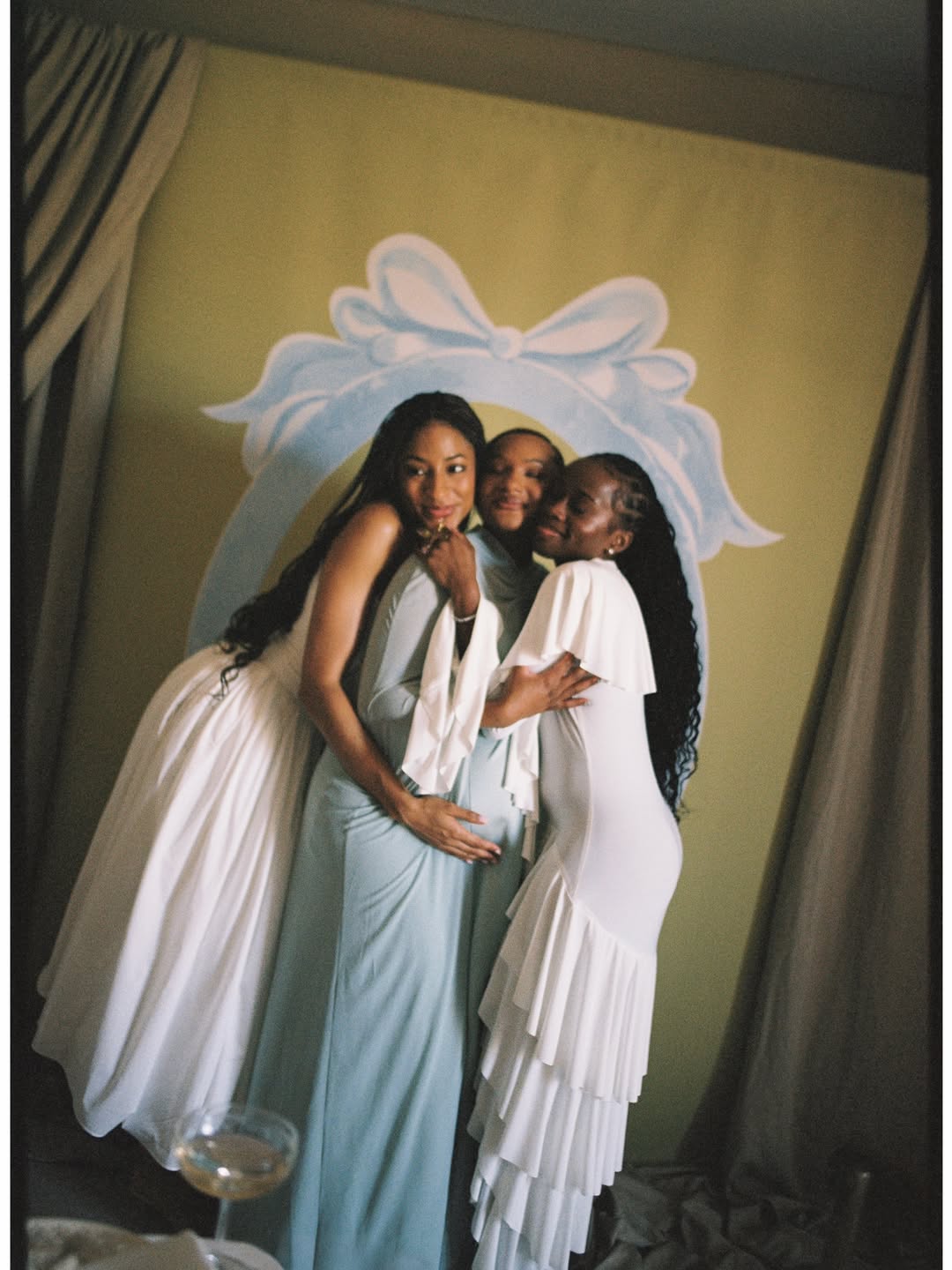 Temi Ajibade in her ice-blue maternity gown sharing a close embrace with two friends dressed in white at her swan-themed baby shower.