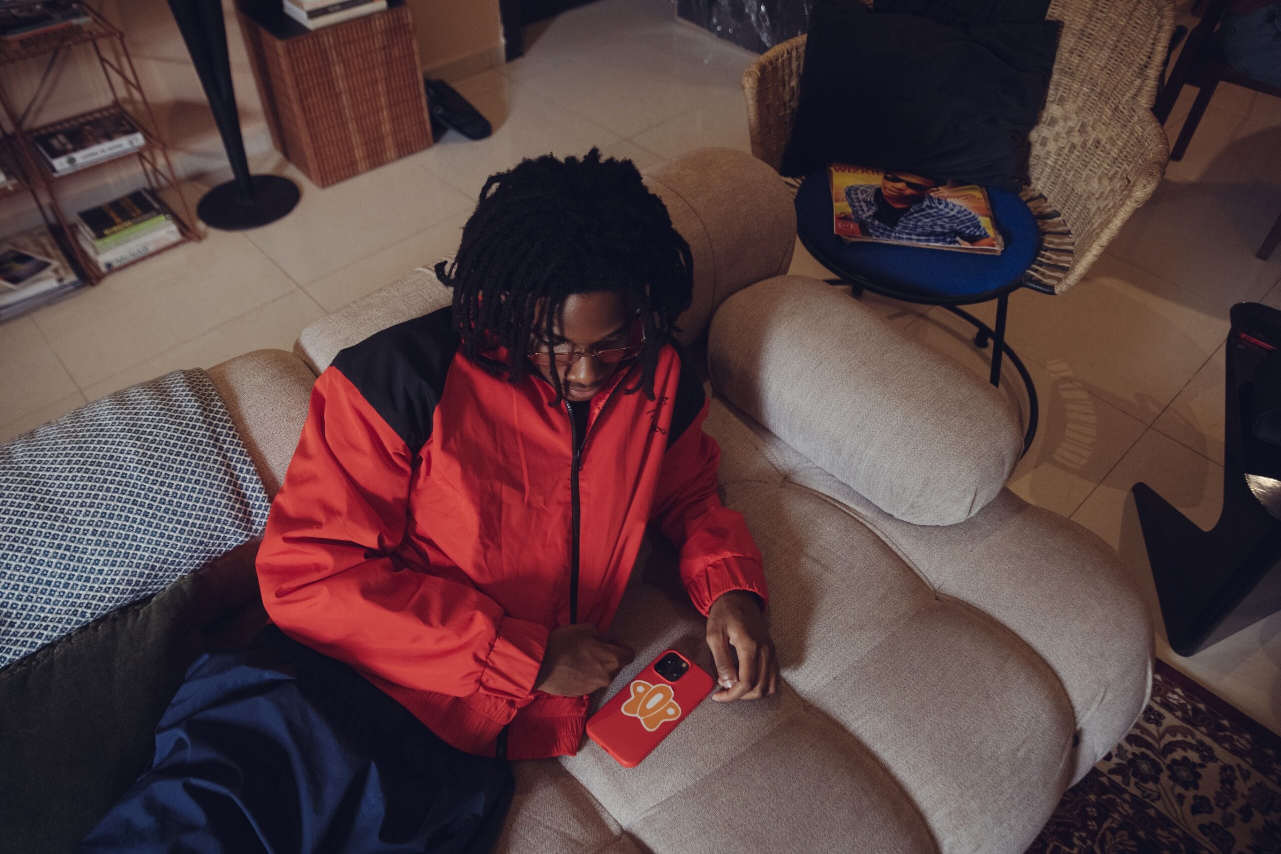 Afrobeats artist Taves looking at his mobile phone while sitting on a couch, with a Wizkid vinyl record visible on a table in the background.
