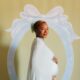 Temi Ajibade cradling her pregnancy baby bump while wearing a mint-tinted ice blue silk maternity gown with a matching cape at her swan-themed baby shower.