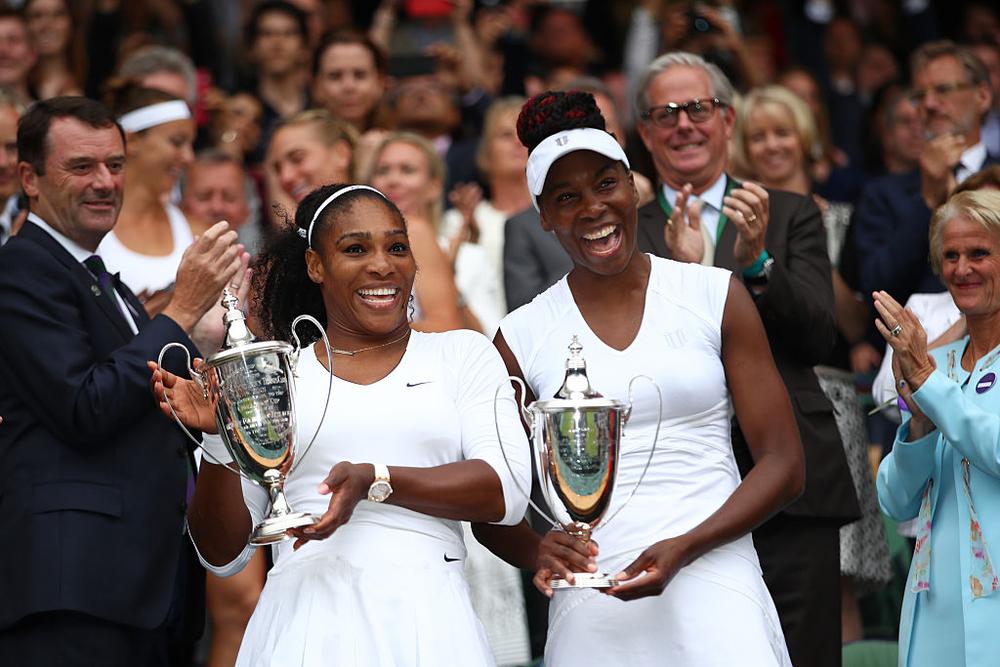 Serena and Venus Williams laughing on the grass court while holding up their matching silver Wimbledon women's doubles championship trophies.