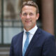 A professional portrait of Peter Vowles, the newly appointed British High Commissioner to Nigeria, wearing a navy blue suit and patterned tie, smiling outdoors.